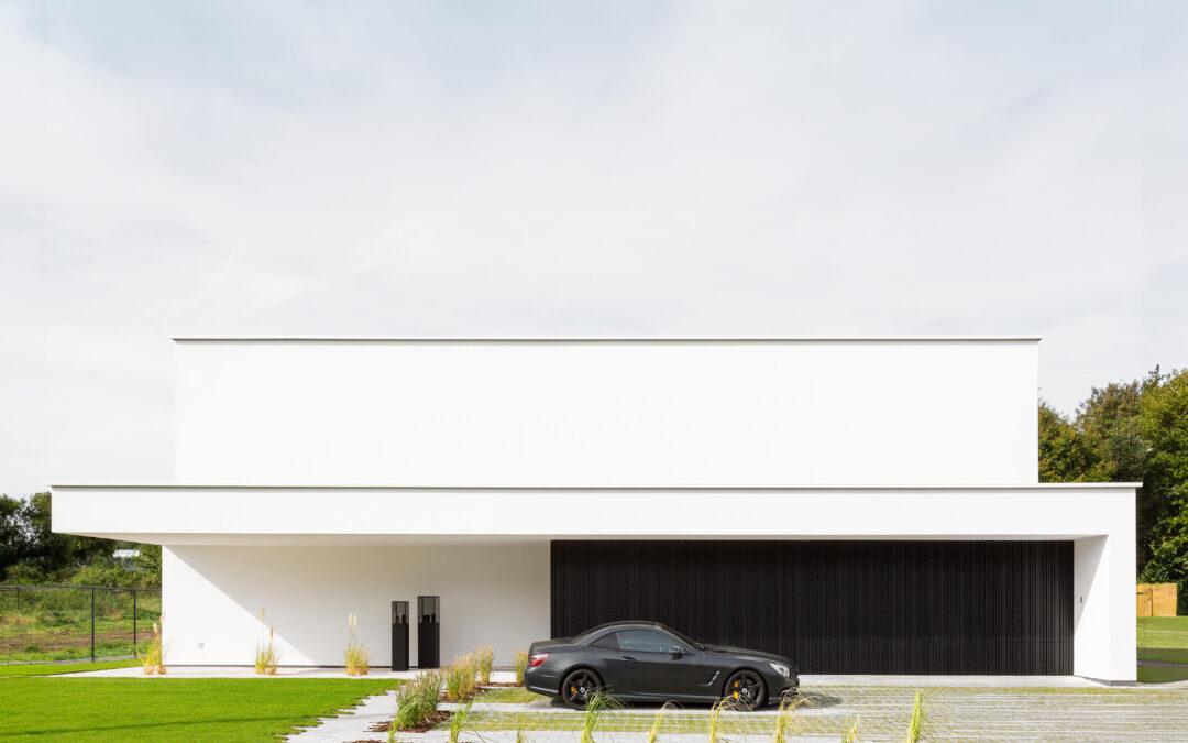 Strakke witte woning met zwarte garagepoort, ontworpen door Jonathan Coenen, met aangelegde tuin, oprit en parkeerplaats