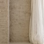 Detail van Mineral Touch gietvloer tegen ruwe bakstenen muur met lichte gordijnen in loftinterieur.