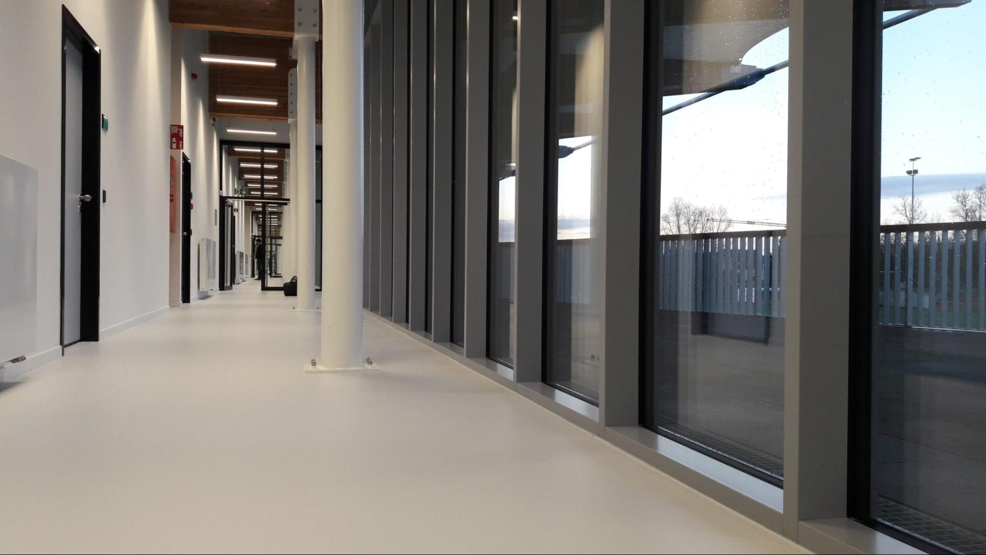 Epoxy gietvloer grijs Moderne gang in kantoorgebouw met naadloze epoxy vloer en strakke afwerking.<br />