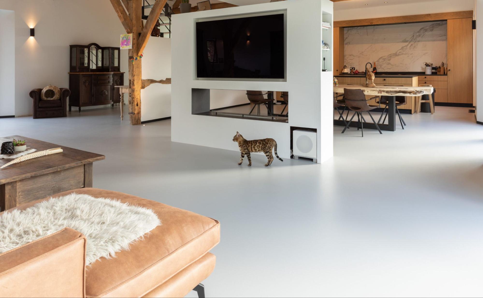 Grijze PU gietvloer die doorloopt van living naar open keuken in een gerenoveerde landelijke woning met houten balken.<br />
